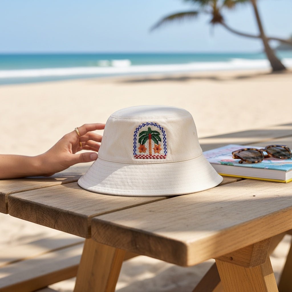 JOYCO Palm Mirage beige bucket hat with colorful palm tree embroidery, resting on a wooden table at the beach.