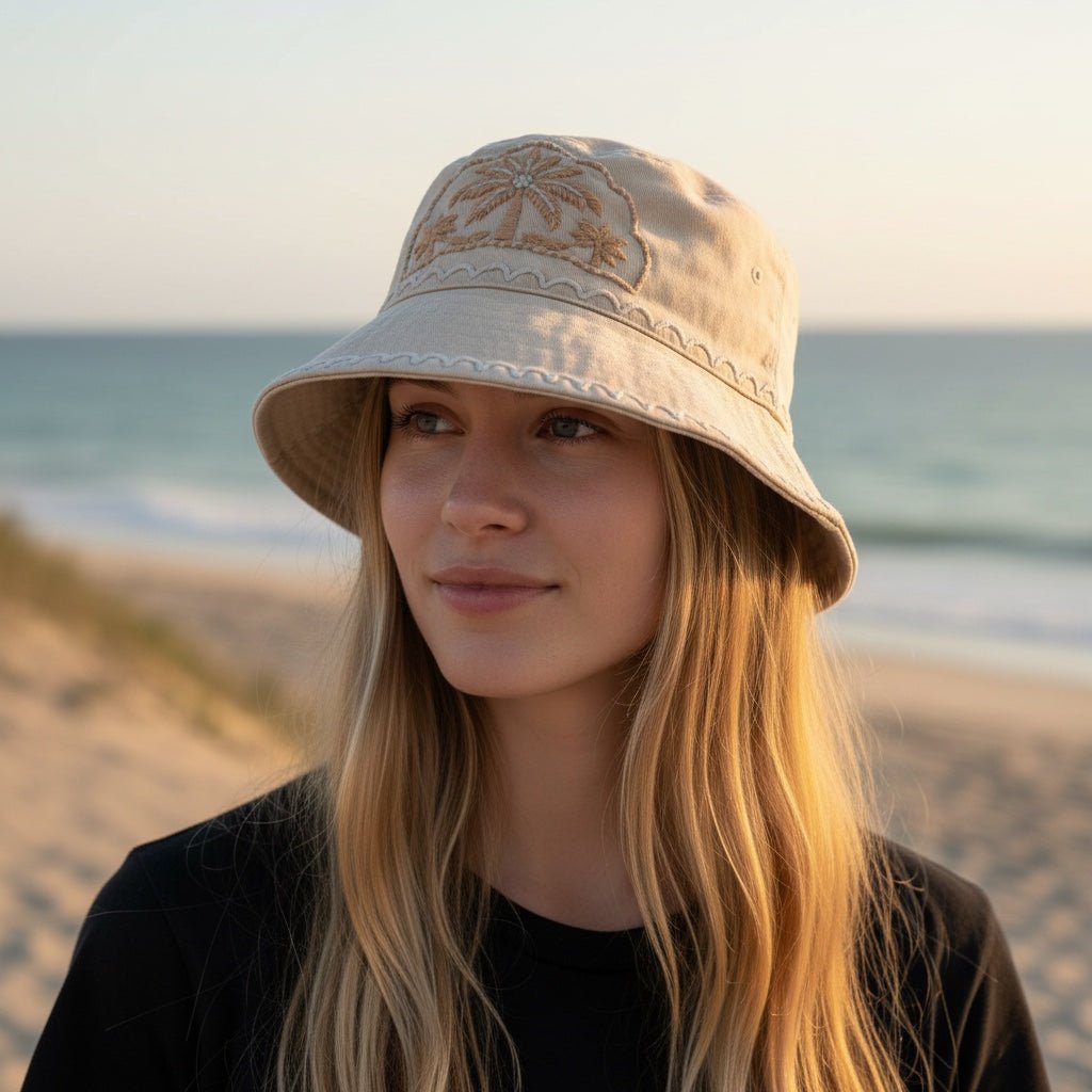 A woman wearing a beige JOYCO bucket hat with tropical palm tree embroidery on a sunny beach at sunset.