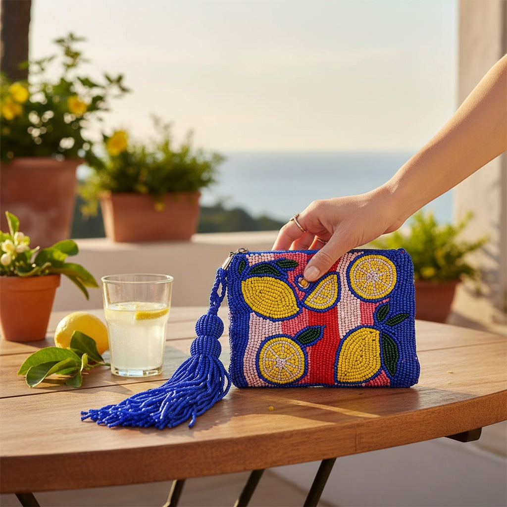 JOYCO Capri Lemon embroidered pouch with vibrant beaded fruit design, blue tassel, and zip closure on a patio table.