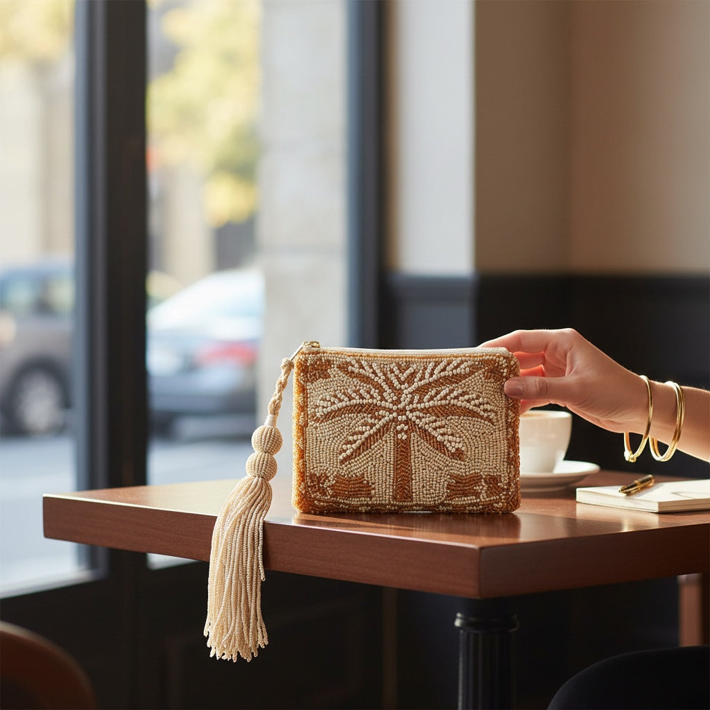 JOYCO Palm Glow beige pouch with shimmering gold palm tree embroidery and a long beaded tassel on a cafe table