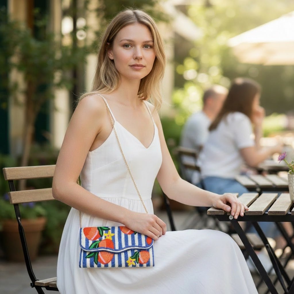 JOYCO Peach Riviera embroidered clutch with blue stripes and peach motifs, worn by a woman in a white dress at a cafe.