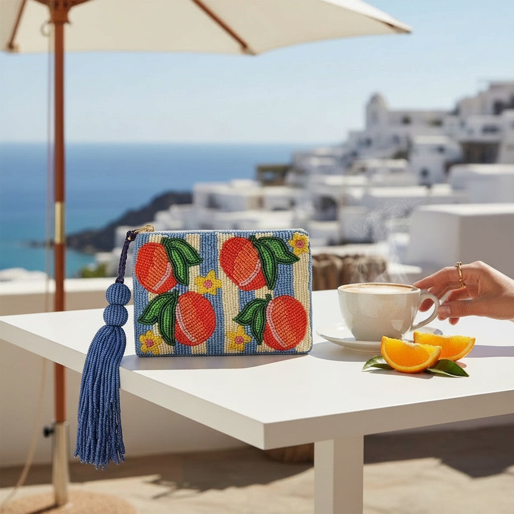 JOYCO Peach Riviera embroidered pouch with blue tassel, featuring peach motifs on a striped background on a cafe table