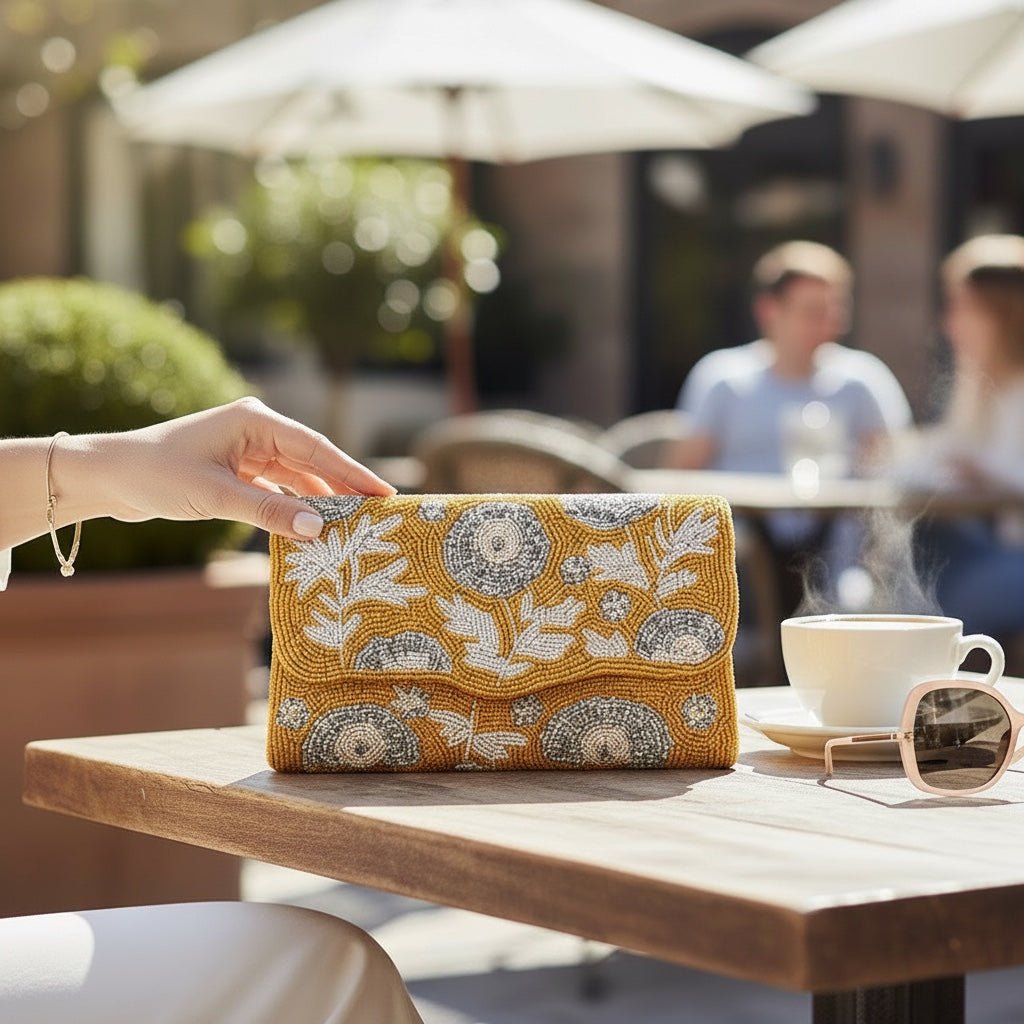 JOYCO Soleil Bloom mustard yellow clutch with intricate silver and white floral embroidery on a cafe table.